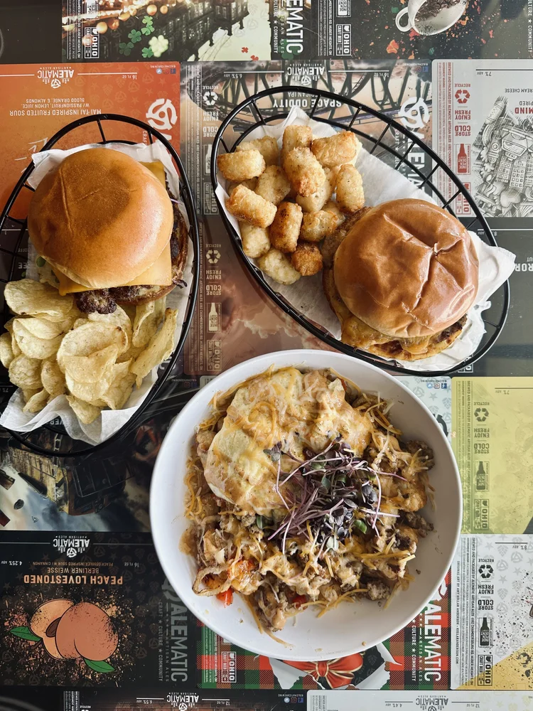Burgers, tots, chips, and a loaded bowl on Alematic table paper.